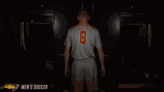 a man stands in a dark room with his arms crossed and a men 's soccer logo in the corner