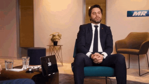 a man in a suit and tie sits in a chair with his eyes closed in front of a sign that says mg