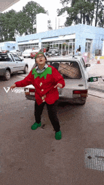 a person in a red elf costume is dancing in front of a silver car with a sticker that says viggle ai