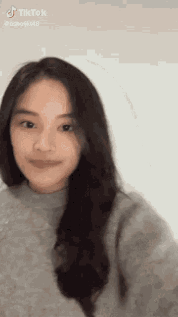 a woman with long black hair is wearing a grey sweater and taking a selfie on tiktok .