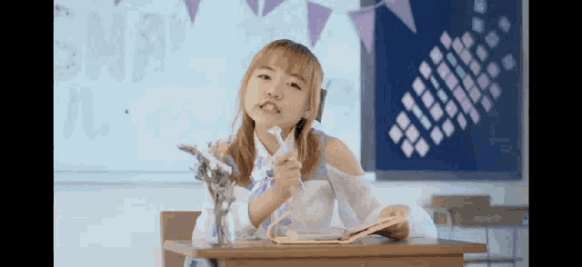 a young girl is sitting at a desk in a classroom holding a book and a pen .