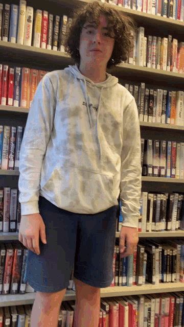 a man in a tie dye hoodie stands in front of a bookshelf filled with books