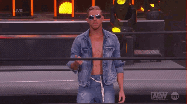 a man wearing sunglasses and a denim jacket is standing in front of a fence that says aew on it