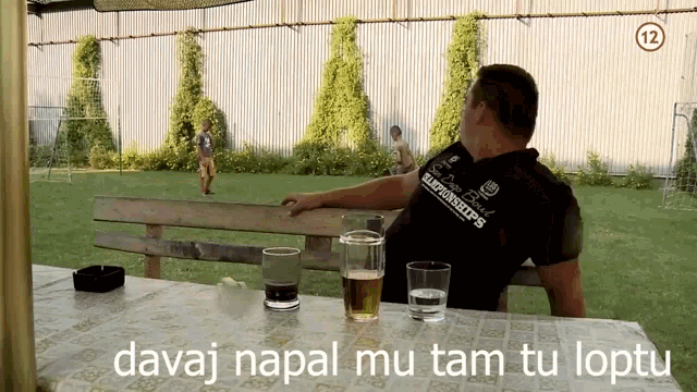 a man sits at a table with two glasses of beer and the words " davaj napal mu tam tu loptu " above him