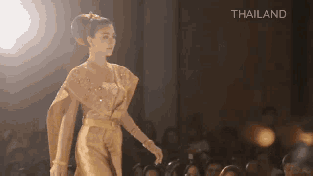 a woman in a gold dress walks down a runway in front of a crowd with the word thailand below her