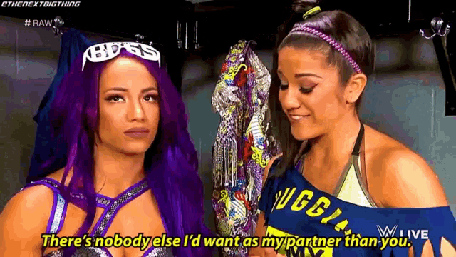 two female wrestlers are talking to each other in a locker room . one of the wrestlers has purple hair .