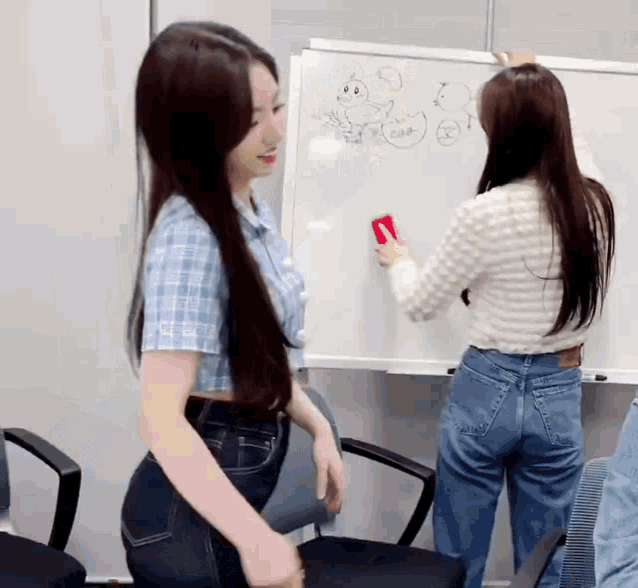 two women are standing in front of a whiteboard and one is holding a red marker .