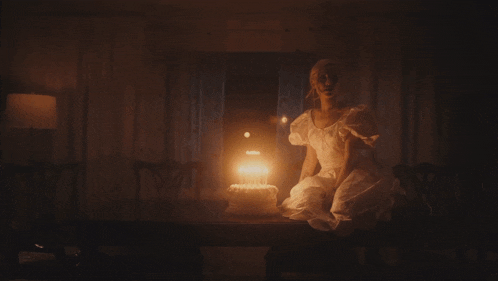a woman in a white dress sits at a table with a birthday cake with candles on it