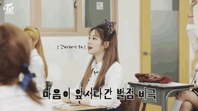 a girl with a bow in her hair is sitting at a desk in a classroom with a twice logo in the corner