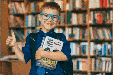 a young boy wearing glasses and a backpack holds a book in front of a bookshelf and gives a thumbs up sign