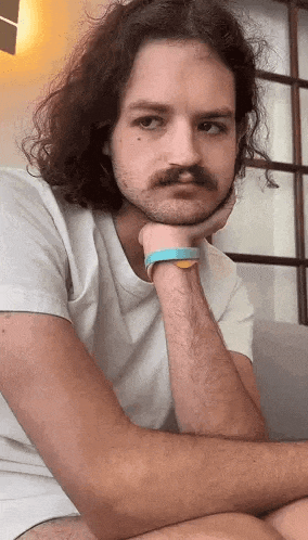 a man with long curly hair and a mustache is sitting with his hand on his chin .