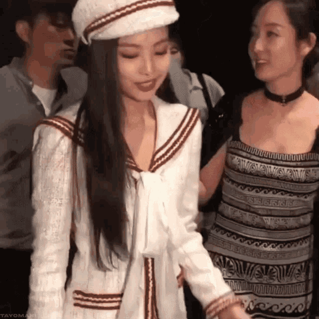 a woman wearing a hat and a white jacket stands next to a woman in a dress .