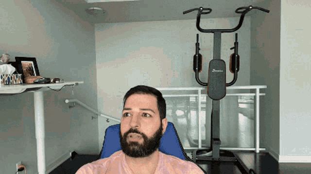 a man with a beard sits in a chair in front of a gym machine that says ' stamina ' on it