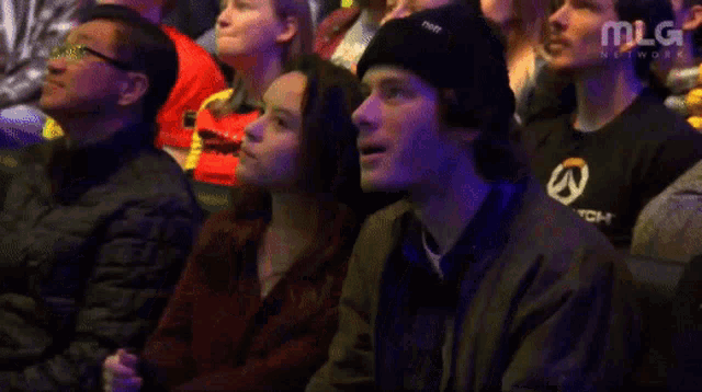 a man in a black overwatch shirt sits in a crowd of people