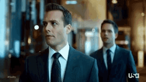 two men in suits and ties are walking down a hallway in a building .
