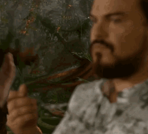 a man with a beard and a white shirt is standing in front of a plant .