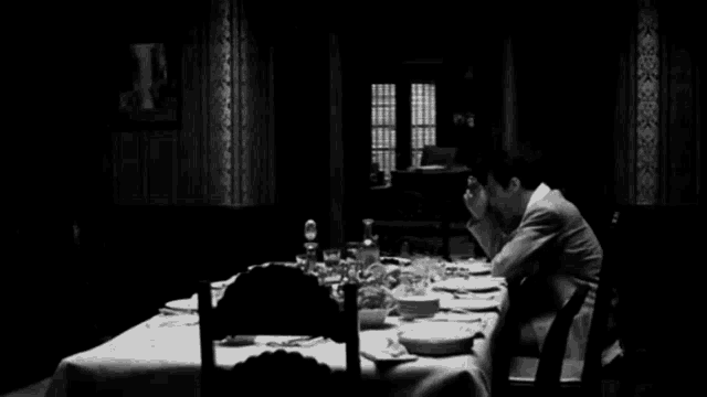 a black and white photo of a man sitting at a long table