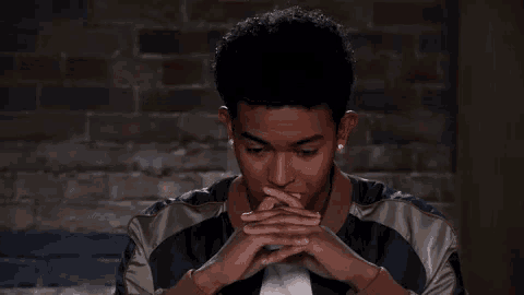 a young man in a jacket is sitting in front of a brick wall with his hands folded .