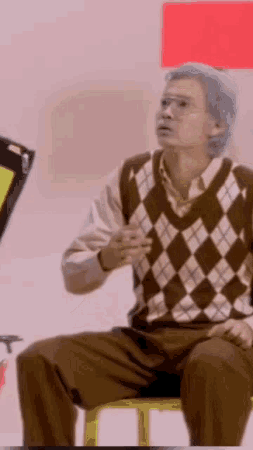 a man with gray hair is sitting on a yellow chair wearing a plaid sweater and glasses .