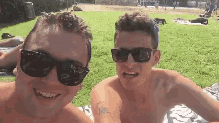 two shirtless men wearing sunglasses are sitting on a blanket in the grass .
