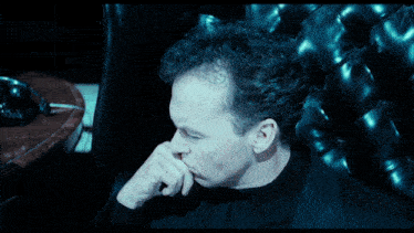 a man in a black suit is sitting in a chair and pointing at something