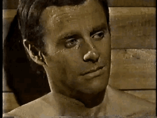 a shirtless man is standing in front of a wooden wall and looking at the camera .