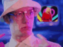 a man wearing a hat and glasses is standing in front of a television .