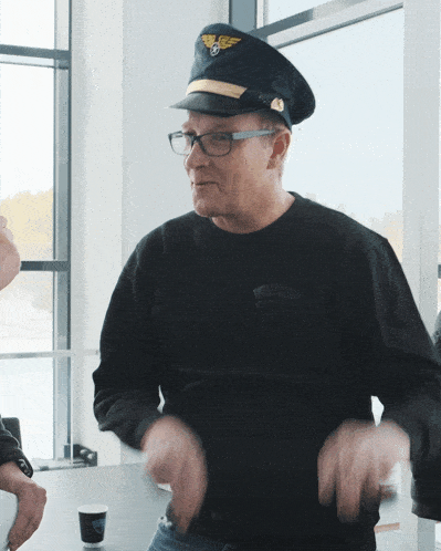 a man wearing a pilot hat and glasses is standing in front of a window