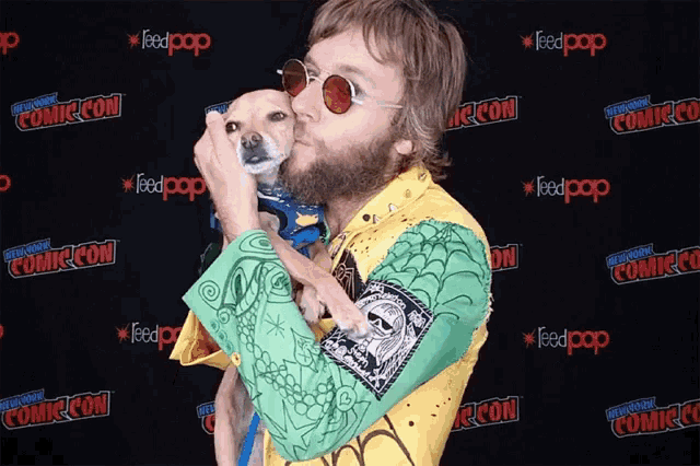 a man is kissing a small dog in front of a comic con backdrop