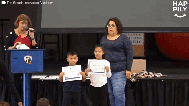 a woman stands at a podium with two children holding certificates in front of a large screen that says hap pily