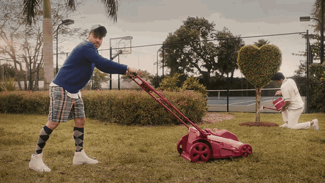 a man pushes a pink lawn mower while a man waters a heart shaped tree
