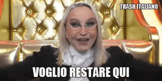 a woman wearing glasses is sitting on a gold couch and says " voglio restare qui " .