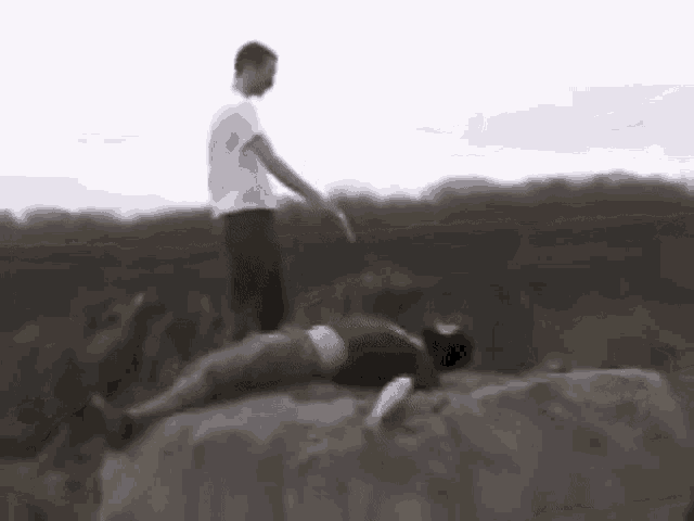 a man is standing next to a woman laying on the ground on a rock .