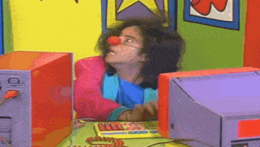 a clown with a red nose is sitting at a desk with a computer .