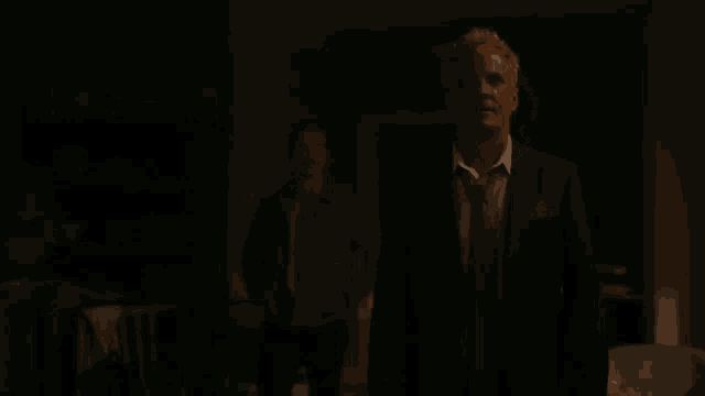 a man in a suit and tie is standing in a dark room talking to another man .