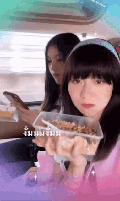 two girls are sitting in a car eating food and one of them is looking at her phone .