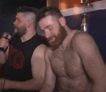 a shirtless man with a beard is talking into a microphone while another shirtless man looks on