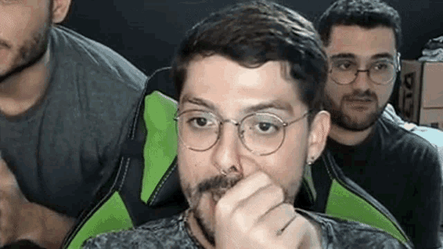 a man with glasses and a beard is covering his nose while sitting in a chair .