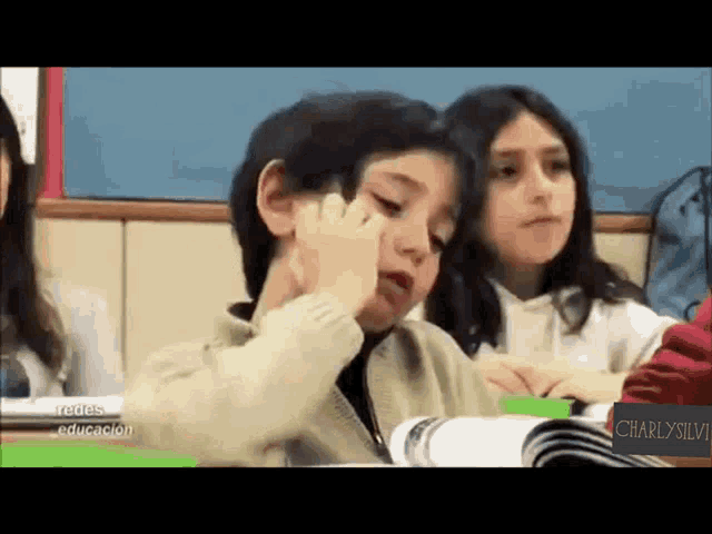 a group of children are sitting in a classroom and one of them is covering his eyes with his hand .