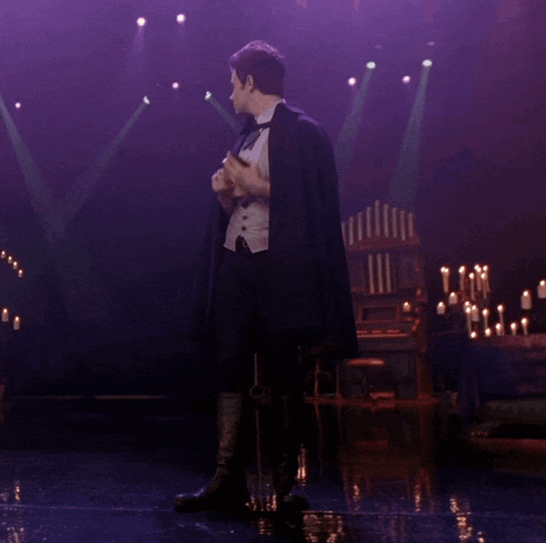 a man in a tuxedo and cape stands on a stage with candles in the background
