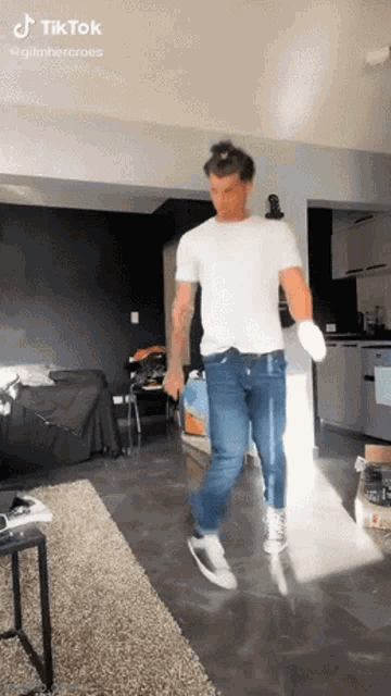 a man in a white shirt and blue jeans is dancing in a living room with tiktok written on the bottom