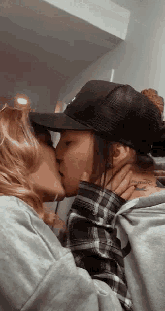 a woman with a tattoo on her neck kisses another woman with a hat on