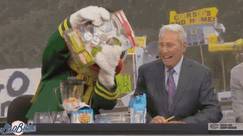 a man in a suit and tie is sitting at a table with a mascot holding a blender full of cereal