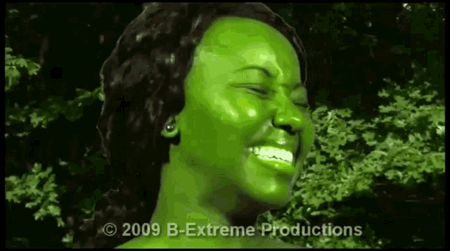 a woman with green paint on her face is smiling in front of a forest .