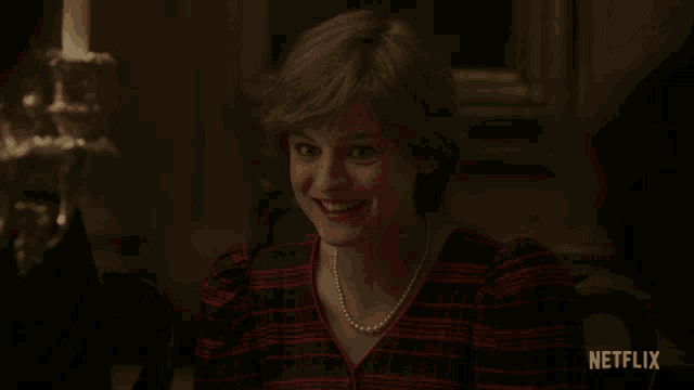 a woman wearing a plaid shirt and a pearl necklace is sitting at a table with a netflix logo behind her