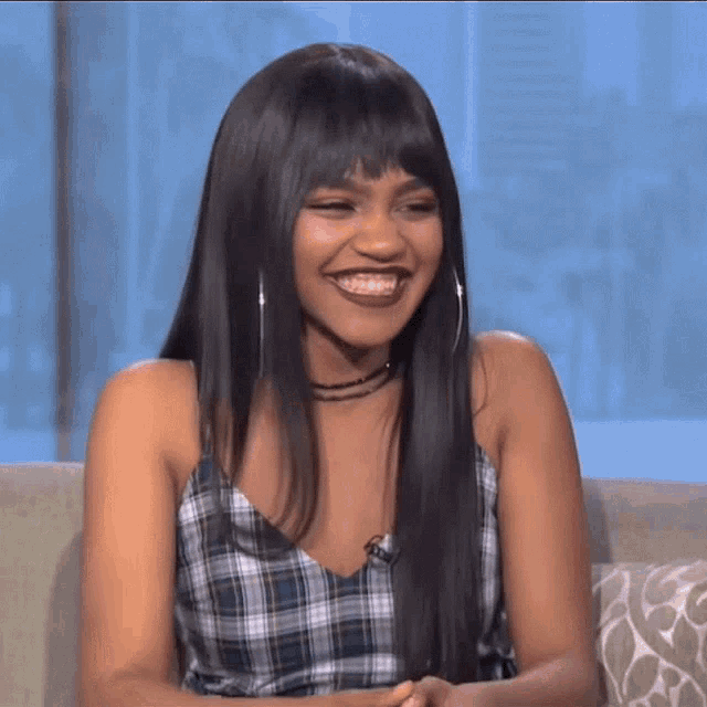 a woman wearing a plaid top and hoop earrings smiles while sitting on a couch