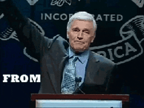 a man in a suit and tie stands at a podium with his arms in the air and the words from behind him