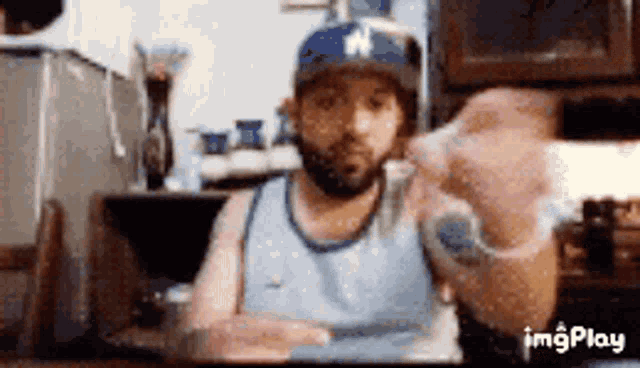 a man with a beard wearing a baseball cap and a blue tank top is sitting at a table .