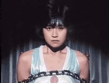 a woman in a blue tank top is chained to a chair and looking at the camera .