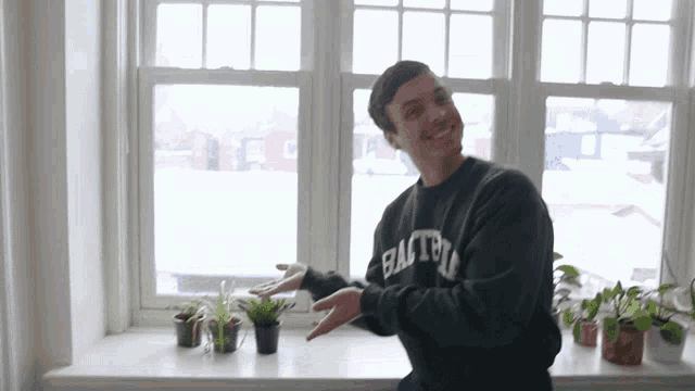 a man wearing a green easton sweatshirt stands in front of a window with potted plants